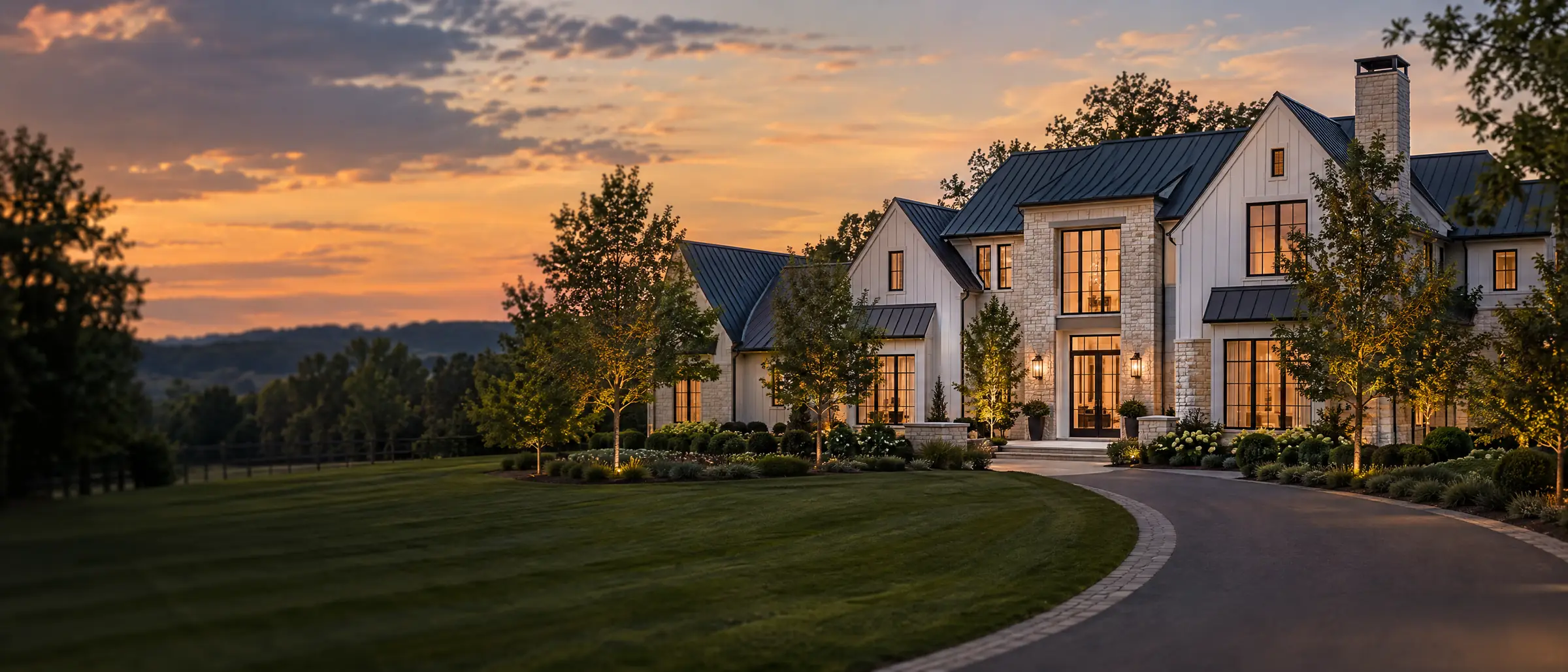 A finished custom estate home at dusk, cream stone and white board with warm light in tall black windows