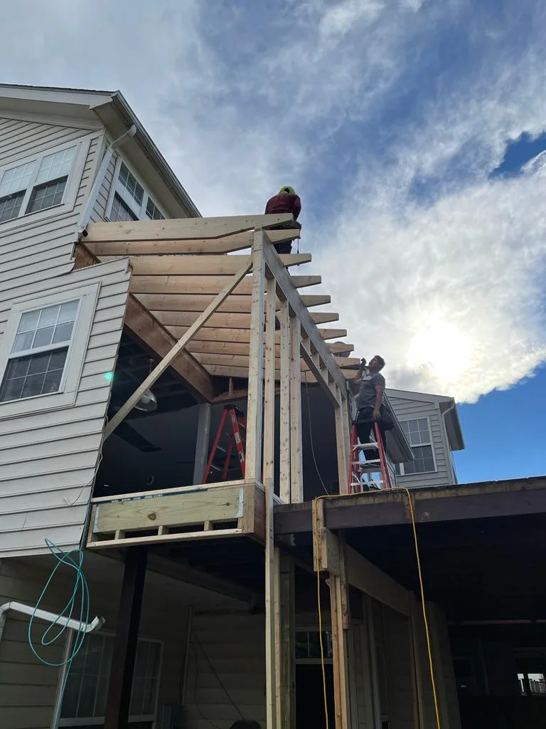Roof rafters of a new porch structure silhouetted against a bright sky, with a worker on a ladder.