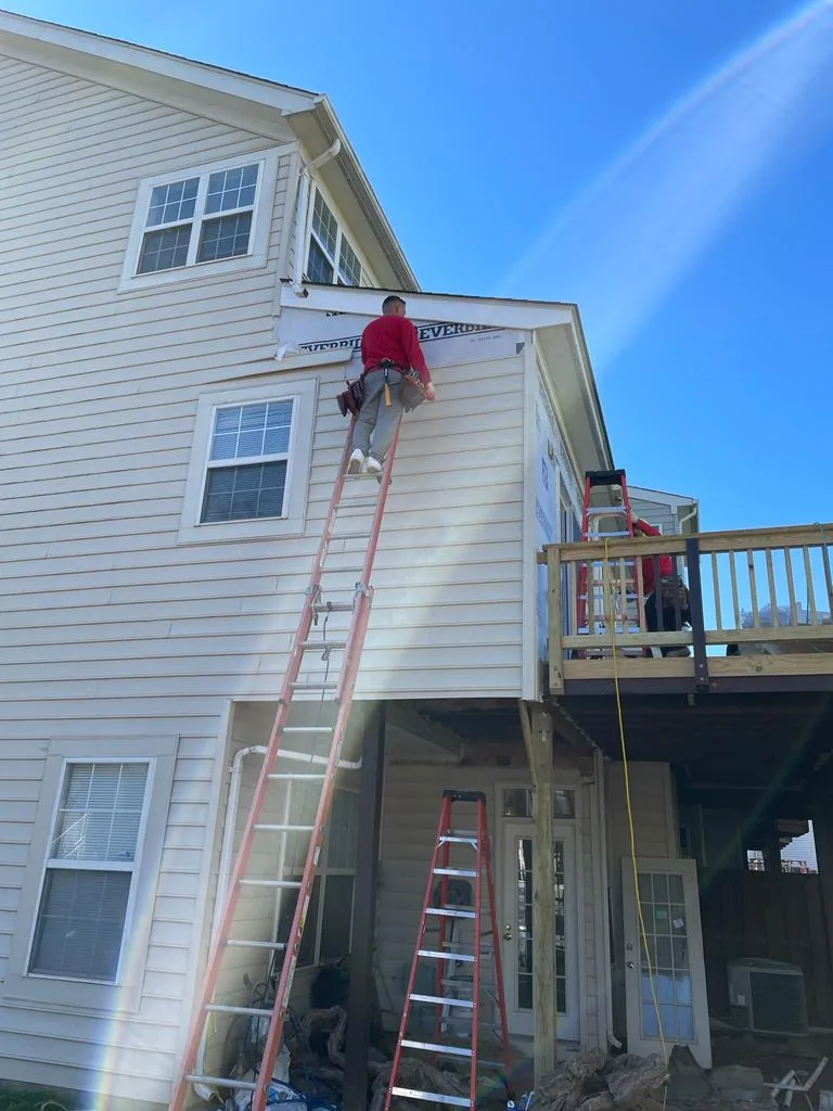 A worker high on an extension ladder replacing siding on the top floor of a house above a deck.