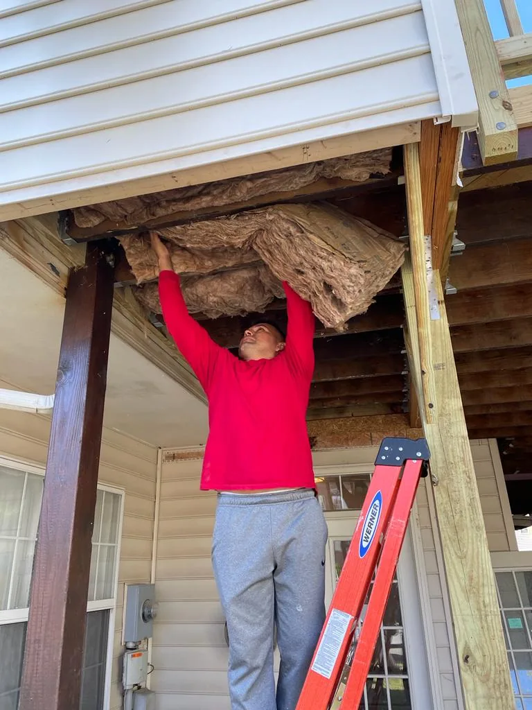A worker lifting a batt of insulation into the ceiling of a covered porch.